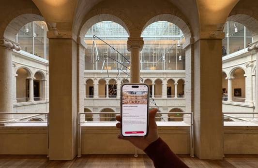 An arm reaching out holding a cell phone with the Harvard Art Museum home page on the screen, while inside the Harvard Art Museum