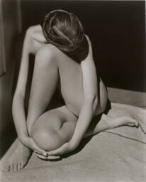 Black-and-white photograph of a seated nude figure curled inward, arms wrapped around legs, head bowed